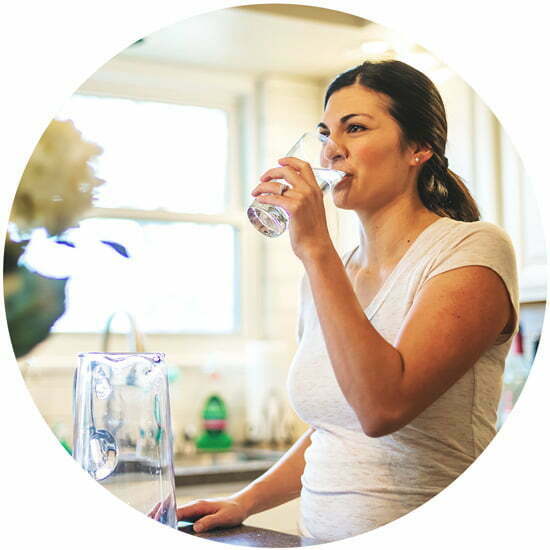 Das Bild zeigt eine Frau, die ein Glas Wasser trinkt. Das Trinken von Wasser ist wichtig für die Unterstützung der Leberentgiftung und die allgemeine Gesundheit. Die Frau trägt ein helles Oberteil und befindet sich in einem Raum mit neutralem Hintergrund.